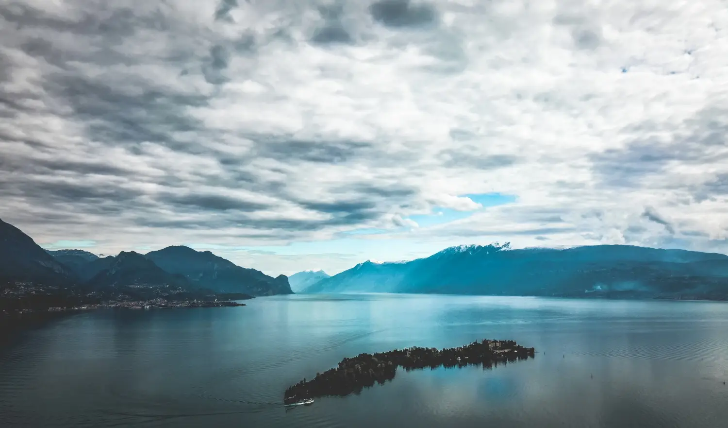 Luftaufnahme des Gardasees mit einer kleinen Insel im Vordergrund, umgeben von Bergen und einem bewölkten Himmel.