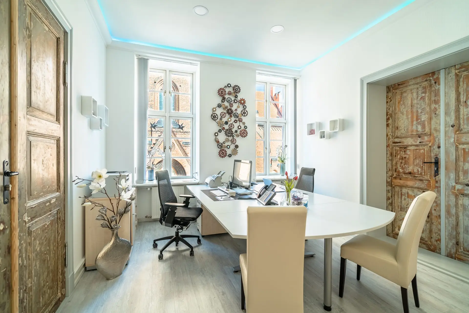 Ein modernes Büro mit weißem Tisch, ergonomischen Stühlen und großen Fenstern, durch die die Fassade eines historischen Gebäudes sichtbar ist. Das Interieur kombiniert Tradition und Moderne.