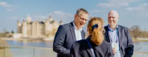 Drei Personen im Business-Outfit stehen auf einer Terrasse und unterhalten sich, im Hintergrund ist das Schweriner Schloss zu sehen.