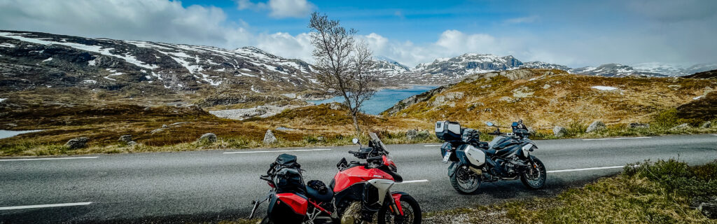 Motorradreise Norwegen – zwei Motorräder auf einer Panoramastraße mit Blick auf schneebedeckte Berge und Fjord