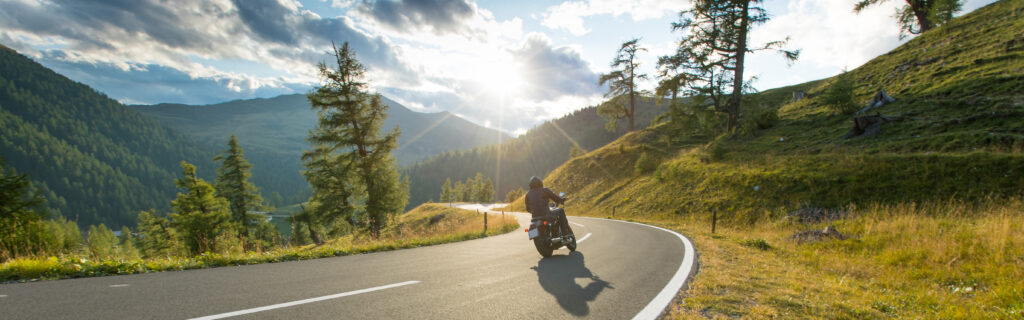 „Motorradreise Alpen – Motorradfahrer auf kurviger Bergstraße bei Sonnenuntergang mit Bergpanorama“