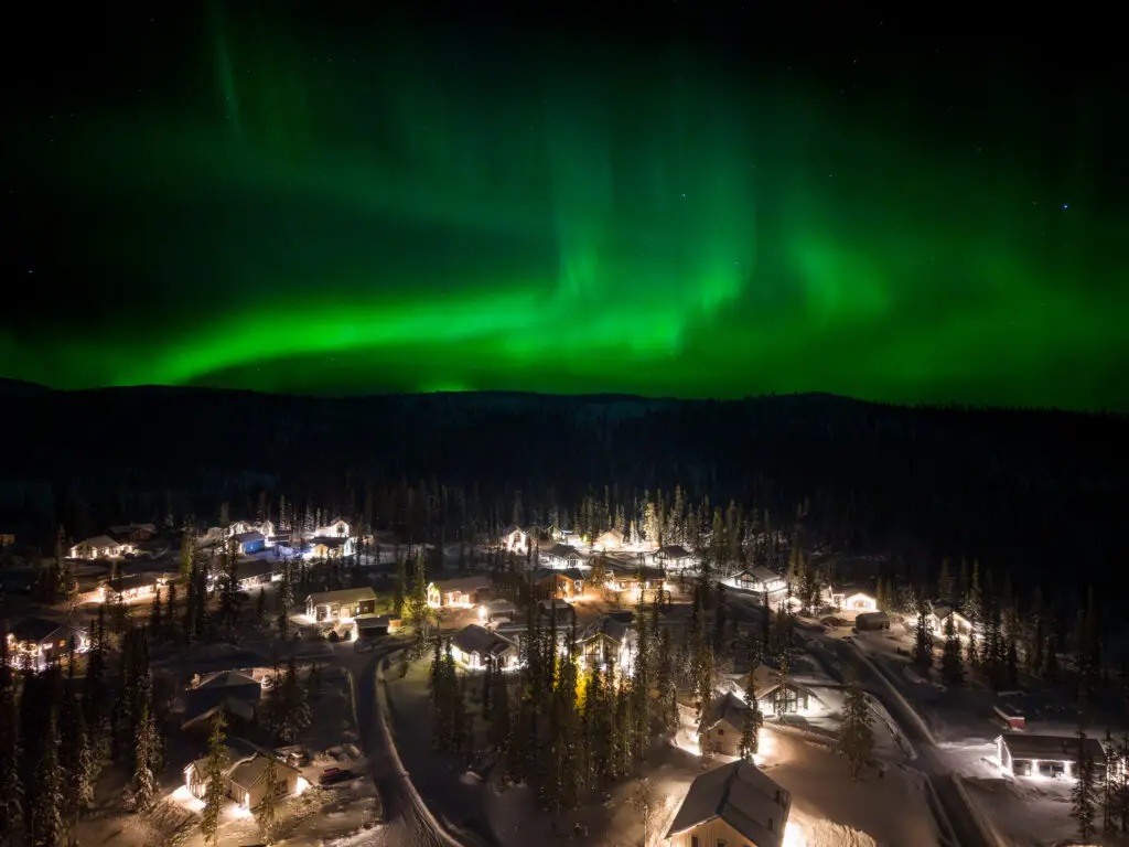 Polarlichter über einem verschneiten Dorf in Lappland, Schweden, mit beleuchteten Holzhäusern und grünem Nachthimmel