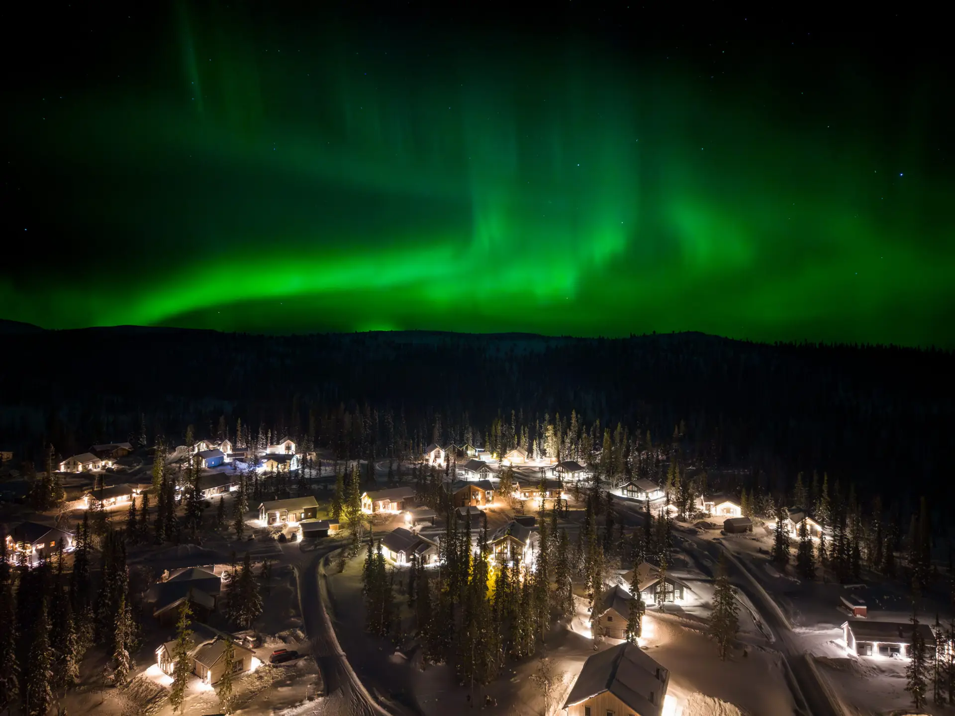 Polarlichter über einem verschneiten Dorf in Lappland, Schweden, mit beleuchteten Holzhäusern und grünem Nachthimmel