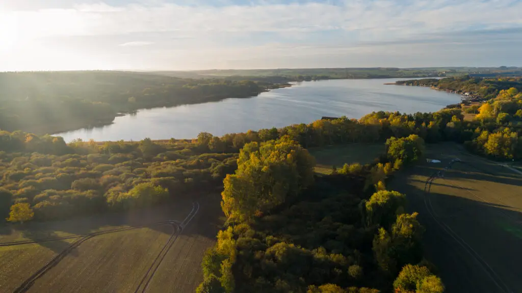 Luftaufnahme des Inselsees in Güstrow mit herbstlich gefärbten Wäldern, angrenzenden Feldern und warmem Sonnenlicht.