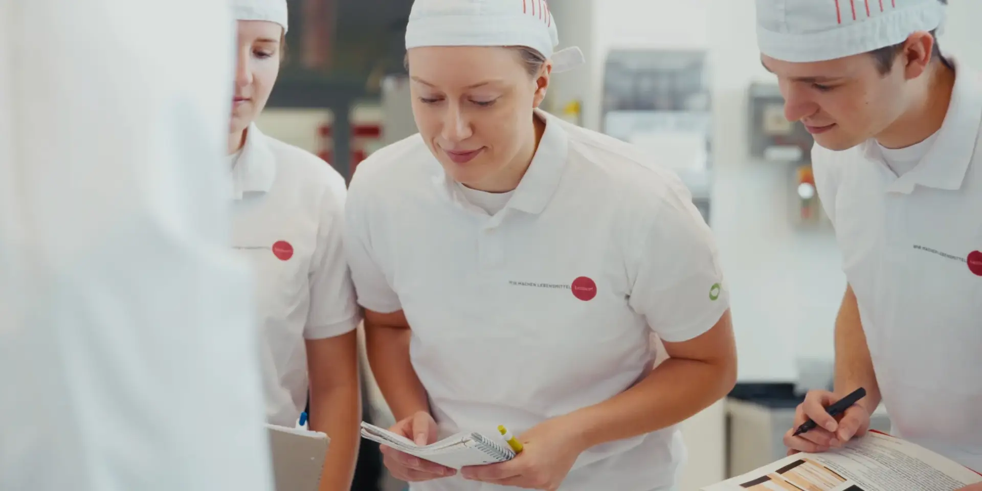 Drei Mitarbeitende in weißer Arbeitskleidung besprechen Unterlagen in einem Lebensmittelbetrieb. Authentische Aufnahme für einen Unternehmensfilm.